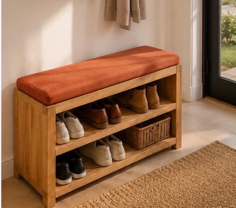 K&ouml;nnte beinhalten: Eine rechteckige Schuhbank aus Holz mit einem Sitzpolster aus rostorangefarbenem Stoff. Das M&ouml;belst&uuml;ck verf&uuml;gt &uuml;ber zwei offene Regalebenen, die mit verschiedenen Paar Schuhen und einem kleinen geflochtenen Korb best&uuml;ckt sind. Der Rahmen ist hell.