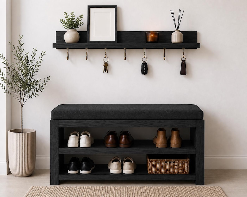 K&ouml;nnte beinhalten: Eine schwarze Flurbank aus Holz mit gepolsterter Sitzfl&auml;che und zwei offenen Regalen f&uuml;r Schuhe und einen Weidenkorb. Dar&uuml;ber befindet sich ein passendes schwarzes Wandregal mit sechs Messinghaken f&uuml;r Schl&uuml;ssel und Dekorationsgegenst&auml;nde.