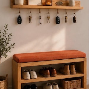 K&ouml;nnte beinhalten: Eine Flurbank aus hellem Holz mit orangefarbenem Polstersitz und zwei Ablagef&auml;chern f&uuml;r Schuhe und einen geflochtenen Korb. Dar&uuml;ber befindet sich ein passendes Wandregal aus Holz mit sieben Metallhaken, an denen Schl&uuml;ssel h&auml;ngen.