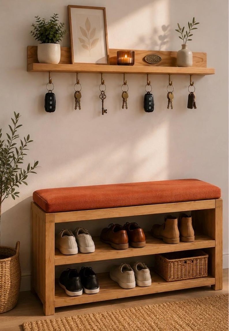 K&ouml;nnte beinhalten: Eine Flurbank aus hellem Holz mit orangefarbenem Polstersitz und zwei Ablagef&auml;chern f&uuml;r Schuhe und einen geflochtenen Korb. Dar&uuml;ber befindet sich ein passendes Wandregal aus Holz mit sieben Metallhaken, an denen Schl&uuml;ssel h&auml;ngen.