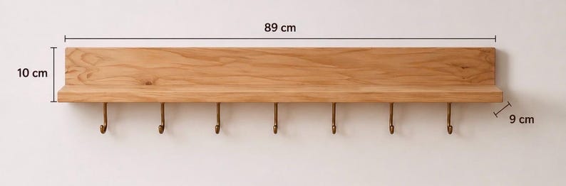 K&ouml;nnte beinhalten: Ein hellbraunes Wandregal aus Holz mit sieben messingfarbenen Metallhaken an der Unterseite. Das Regal ist 89 cm lang, 10 cm hoch und 9 cm tief, wie durch die im Bild sichtbaren Ma&szlig;linien deutlich gekennzeichnet ist.
