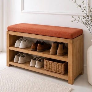 K&ouml;nnte beinhalten: Eine rechteckige Schuhbank aus Holz mit einem leuchtend orangefarbenen Polstersitz. Das M&ouml;belst&uuml;ck verf&uuml;gt &uuml;ber zwei offene Ablagef&auml;cher, in denen sich derzeit mehrere Paar Freizeitschuhe und ein kleiner geflochtener Korb befinden.