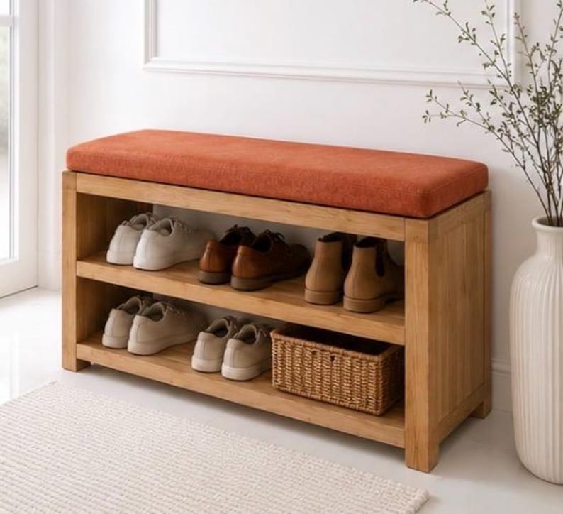 K&ouml;nnte beinhalten: Eine rechteckige Schuhbank aus Holz mit einem leuchtend orangefarbenen Polstersitz. Das M&ouml;belst&uuml;ck verf&uuml;gt &uuml;ber zwei offene Ablagef&auml;cher, in denen sich derzeit mehrere Paar Freizeitschuhe und ein kleiner geflochtener Korb befinden.