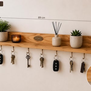 K&ouml;nnte beinhalten: Ein hellbraunes Wandregal aus Holz als Schl&uuml;sselhalter mit sieben messingfarbenen Haken. Das Regal verf&uuml;gt &uuml;ber ein zentrales ovales Schild mit der Aufschrift HOME. Es ist 89 cm lang, 10 cm hoch und 9 cm tief und h&auml;lt verschiedene Schl&uuml;ssel.