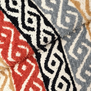 Peut inclure: Une collection de bandes de tissu texturées présentant un motif géométrique répétitif en noir, orange rouille, gris et beige. Chaque bande affiche un design à poils ras surélevés sur une base crème, révélant des formes entrelacées.