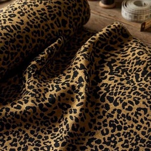 Könnte beinhalten: Eine Stoffrolle mit einem sich wiederholenden schwarzen Leopardenmuster auf beigem Grund. Das Textil ist in weichen Falten drapiert, die die Textur und den Kontrast zwischen den dunklen Flecken und der helleren Grundfarbe des Materials zeigen.