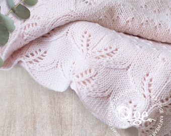 Puede incluir: Una suave manta de bebé de punto rosa con un delicado patrón de encaje. La manta está doblada y drapeada sobre una superficie de madera clara.