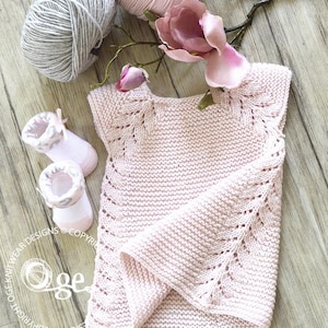 May include: A pink knitted baby sweater with a delicate lace pattern. The sweater has short sleeves and a round neckline. The sweater is laid flat on a wooden surface with a flower and yarn balls.