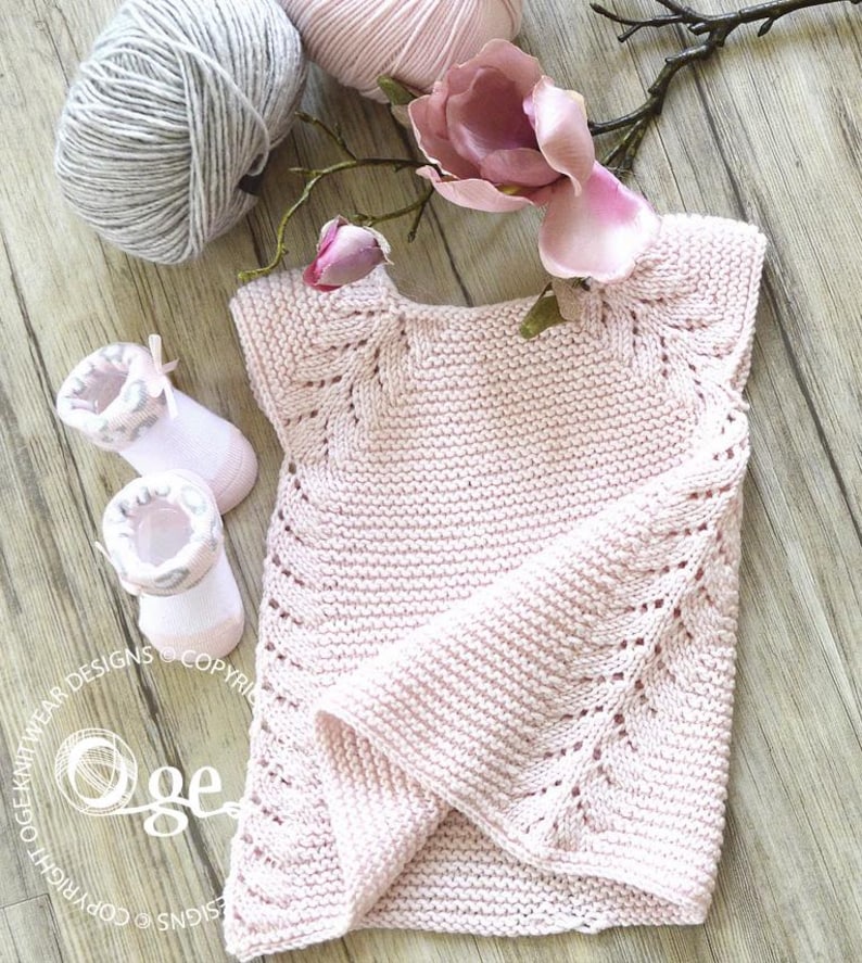 May include: A pink knitted baby sweater with a delicate lace pattern. The sweater has short sleeves and a round neckline. The sweater is laid flat on a wooden surface with a flower and yarn balls.