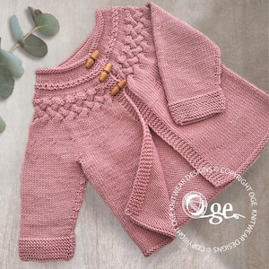 May include: A dusky pink knitted baby outfit, comprising a jacket with wooden toggle buttons and matching trousers. The jacket has a textured cable knit design on the shoulders and sleeves. The set is made of soft, warm yarn.