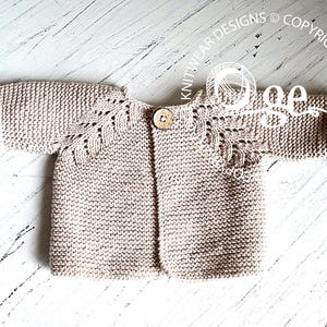 May include: A beige knitted baby cardigan with a button fastening and a delicate lace pattern on the sleeves.