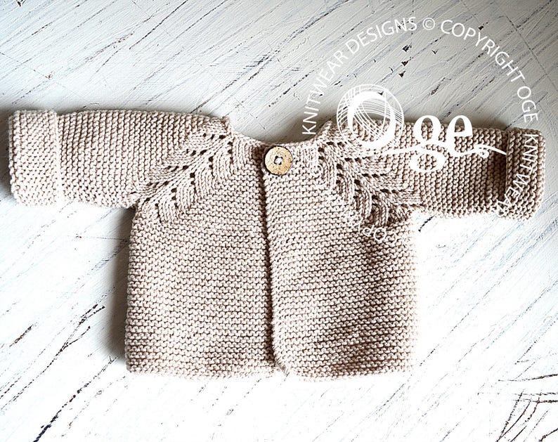May include: A beige knitted baby cardigan with a button fastening and a delicate lace pattern on the sleeves.