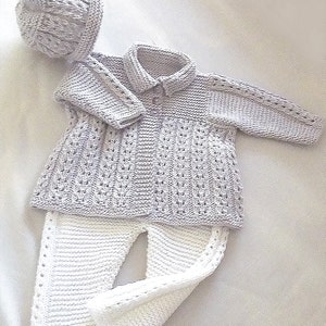 May include: A knitted baby outfit in a light gray color, consisting of a sweater, pants, and a hat. The sweater has a button closure and a ribbed collar. The pants have a straight leg and a ribbed waistband. The hat is a beanie style with a ribbed brim.