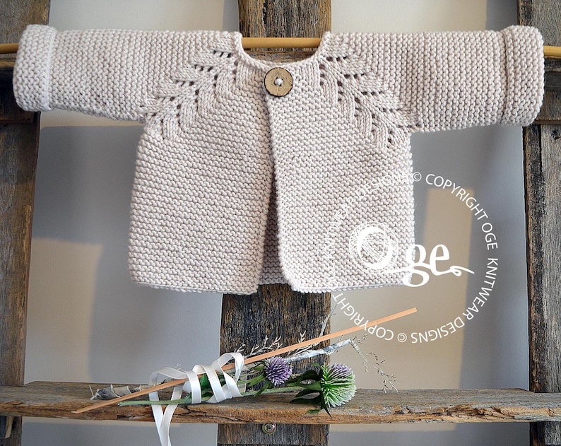 May include: A beige knitted baby cardigan with a button fastening and a delicate leaf pattern on the front. The cardigan is displayed on a wooden shelf.