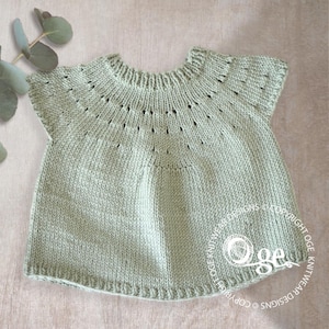 May include: A light green knitted baby top with a delicate lace pattern around the neckline and short sleeves. The top is made with a soft yarn and has a relaxed fit.