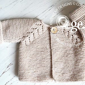 May include: A beige knitted baby cardigan with a button fastening and a delicate lace pattern on the sleeves. The cardigan is laid flat on a white wooden surface.