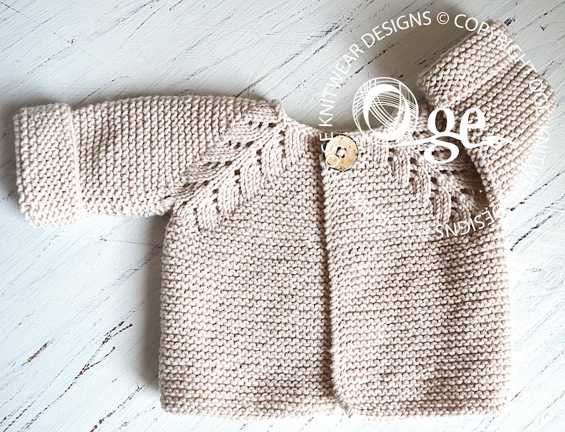 May include: A beige knitted baby cardigan with a button fastening and a delicate lace pattern on the sleeves. The cardigan is laid flat on a white wooden surface.