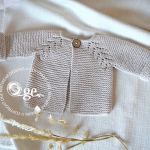 May include: A light beige knitted cardigan with a button fastening and leaf-shaped stitch detail on the shoulders. The cardigan is laid flat on a white fabric background.