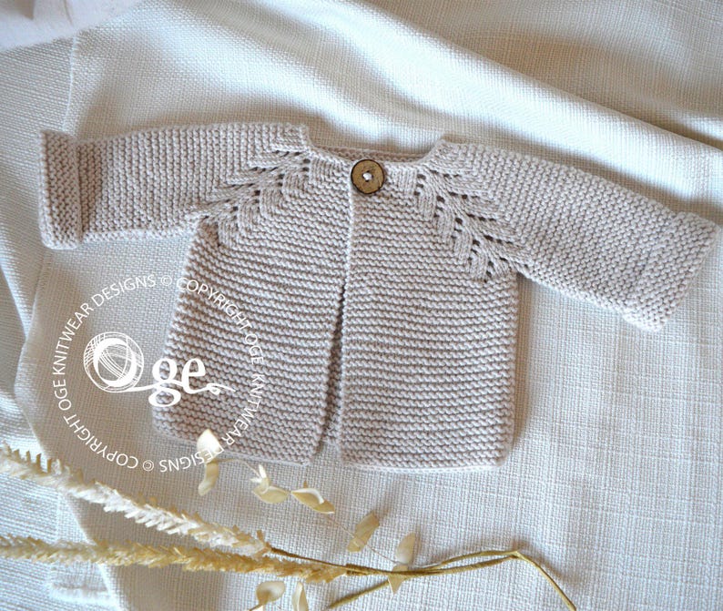 May include: A light beige knitted cardigan with a button fastening and leaf-shaped stitch detail on the shoulders. The cardigan is laid flat on a white fabric background.