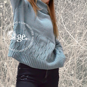 May include: A light blue knitted sweater with a cable knit pattern. The sweater has a high neck and long sleeves. The sweater is worn by a person with dark hair and is photographed against a white background.