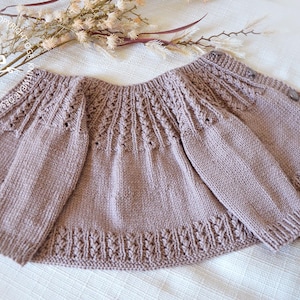 May include: A knitted baby cardigan in a light brown color with a delicate lace pattern. The cardigan has a button closure and long sleeves.