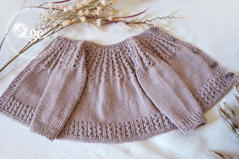 May include: A knitted baby cardigan in a light brown color with a delicate lace pattern. The cardigan has a button closure and long sleeves.
