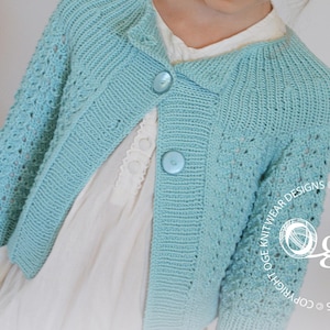 May include: A light blue knitted cardigan with a button closure and a lace pattern. The cardigan is worn over a white dress with lace trim.