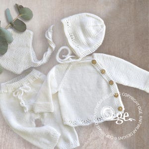 May include: A white knitted baby outfit set including a hat, a cardigan, and pants. The set is made with a delicate stitch pattern and features wooden buttons.