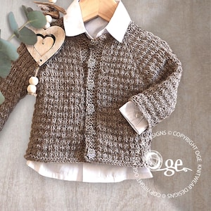 Puede incluir: Un cárdigan de punto marrón con botones con forma de búho, superpuesto a una camisa blanca con cuello. El cárdigan se muestra en una percha de madera, mostrando un diseño de punto texturizado. Una decoración de corazón de madera cuelga a la izquierda.