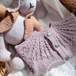 May include: A soft, light purple knitted cardigan with a cable knit pattern and button closures. The cardigan is laid out on a white crocheted blanket with a knitted bunny toy.