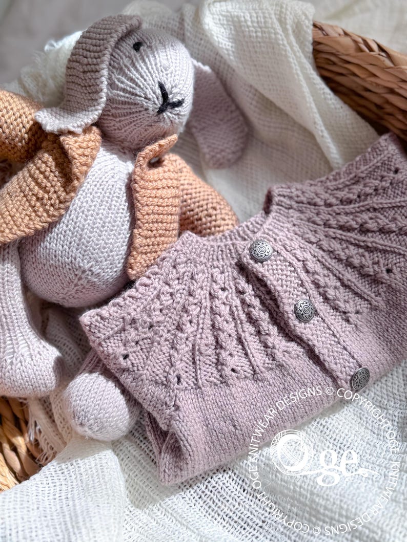 May include: A soft, light purple knitted cardigan with a cable knit pattern and button closures. The cardigan is laid out on a white crocheted blanket with a knitted bunny toy.