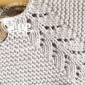 May include: Close-up of a beige knitted fabric with a delicate lace pattern at the neckline. The fabric is soft and textured, with a subtle sheen.