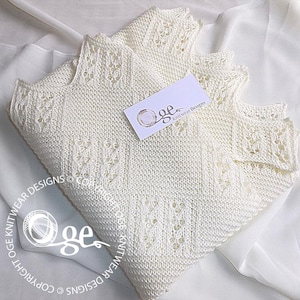 Könnte beinhalten: Eine weiße gestrickte Babydecke mit einem zarten Spitzenmuster. Die Decke ist in der Hälfte gefaltet und hat eine kleine weiße Karte mit dem Text "Oge Knitwear Designs" darauf.