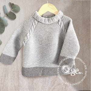 May include: A light grey knitted jumper featuring a ribbed crew neckline, long sleeves with darker grey cuffs, and a matching darker grey hem. The garment includes a textured cable knit pattern along the raglan sleeve seams and a garter stitch body.