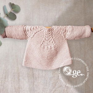 Puede incluir: Un suéter de bebé tejido en rosa claro con intrincados detalles de encaje alrededor del escote. El suéter se muestra en una barra de madera. Las mangas son largas y el diseño es delicado y hecho a mano.