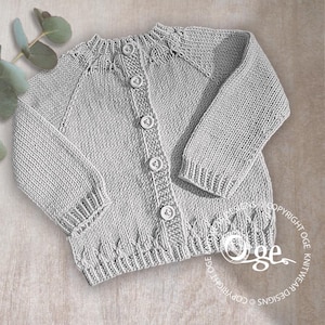 Puede incluir: Un cárdigan de bebé tejido de color gris claro con cierre de botones. El suéter tiene mangas largas y dobladillo y puños acanalados. Los botones son blancos. El cárdigan se muestra sobre una superficie marrón claro.