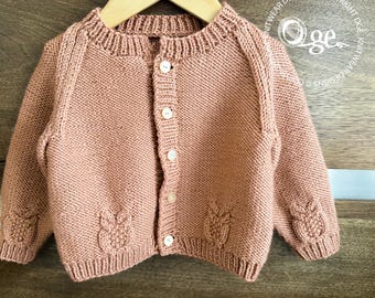 May include: A dusty rose knitted cardigan featuring a ribbed crew neckline, long sleeves, and a front button closure with five white buttons. The garment displays a textured owl motif pattern on the lower front and sleeves, hanging on a hanger.