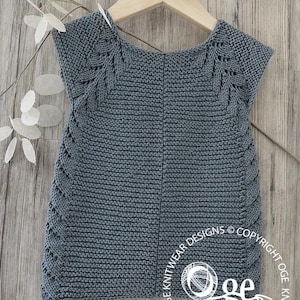 May include: A gray knitted sleeveless top with a lace pattern around the neckline and armholes. The top is hanging on a wooden hanger against a woodgrain background.