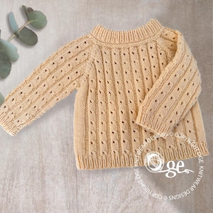 Puede incluir: Un suéter de punto beige claro con una textura acanalada y pequeños detalles de ojal. La prenda de manga larga tiene cuello redondo y se muestra sobre una superficie neutra. El suéter está diseñado para bebés o niños pequeños.