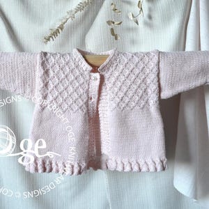 May include: A pink knitted baby cardigan with a diamond pattern on the front and button closure. The cardigan has a ruffled edge and is tied with a ribbon at the shoulders.