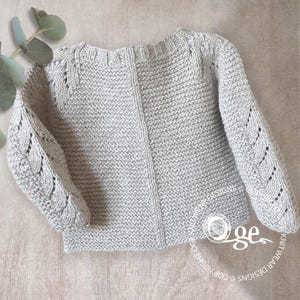 May include: A light gray long-sleeved knit sweater featuring a textured garter stitch body and intricate cable knit patterns running down the center of each sleeve. The garment has a round neckline and a vertical seam detail down the front.
