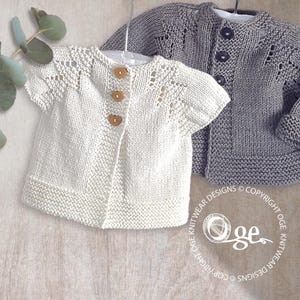 Op de afbeelding: Twee gebreide babyvestjes. Eén is gebroken wit met houten knopen en korte mouwen. De andere is grijs met lange mouwen en donkere knopen. Beide worden op hangers getoond.