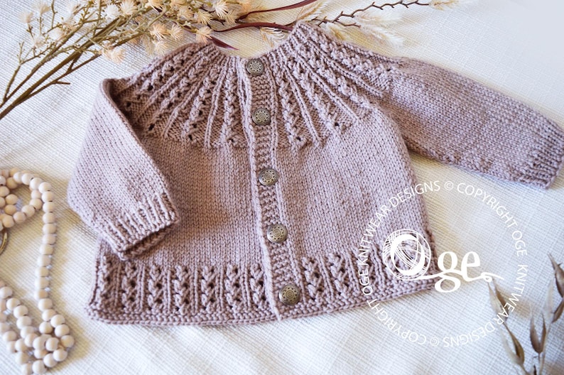 May include: A light brown knitted baby cardigan with a button closure and a delicate lace pattern. The cardigan has a round neckline and long sleeves.
