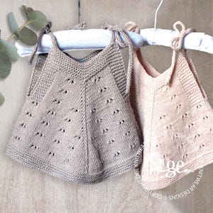 Könnte beinhalten: Zwei gestrickte Baby-Oberteile ohne Ärmel mit einem zarten Spitzenmuster. Das Oberteil links ist hellbraun und das Oberteil rechts ist hellpfirsichfarben. Beide Oberteile haben dünne Träger, die an den Schultern gebunden werden.