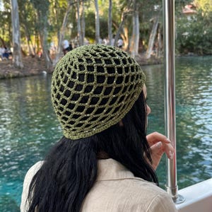 Puede incluir: Un gorro de ganchillo color verde oliva con un patrón de malla abierta en forma de diamante. El hilo está tejido con pequeñas lentejuelas plateadas reflectantes en toda la estructura, creando un brillo sutil en la superficie del gorro.