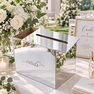 May include: A rectangular mirrored card box with a reflective surface and a top slot for inserting envelopes. The front panel features the word cards written in a cursive script. The box has a clean, modern design with sharp edges and a polished finish.