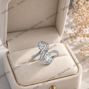 May include: A silver-toned ring featuring an S-shaped bypass design adorned with two prominent round-cut clear stones and smaller pavé-set stones along the band, presented inside a beige velvet-lined jewelry box with a gold-toned clasp.
