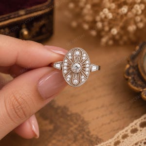 May include: An oval-shaped ring featuring a central round brilliant-cut diamond surrounded by a decorative metal frame with milgrain detailing and smaller round accent stones. The band includes two tapered baguette-cut diamonds on the sides.