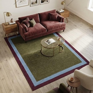 May include: A rectangular area rug featuring a large olive green center panel, a light blue border, and a dark burgundy outer edge. The rug is positioned on a light wood floor beneath a velvet sofa, two round glass coffee tables, and armchairs.