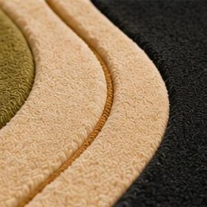 May include: A textured rug featuring curved, layered sections in olive green, cream, and black. The surface has a dense, looped pile construction with distinct raised edges that define the flowing, organic shapes across the fabric material.